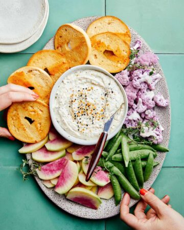 Everything seasoning and caramelized onion dip on a platter with bagels and veggies.