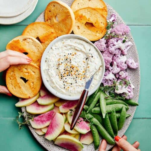 Everything seasoning and caramelized onion dip on a platter with bagels and veggies.