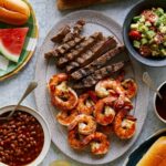Grilled shrimp and steak platter with sides of salad, beans, and watermelon.