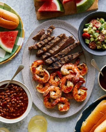 Grilled shrimp and steak platter with sides of salad, beans, and watermelon.