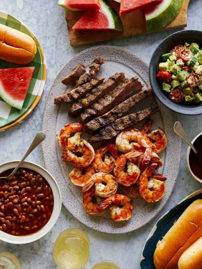 Grilled shrimp and steak platter with sides of salad, beans, and watermelon.