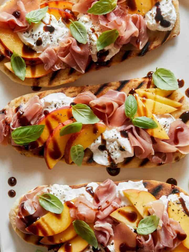 A close up of grilled flatbreads topped with peach, prosciutto, and burrata.