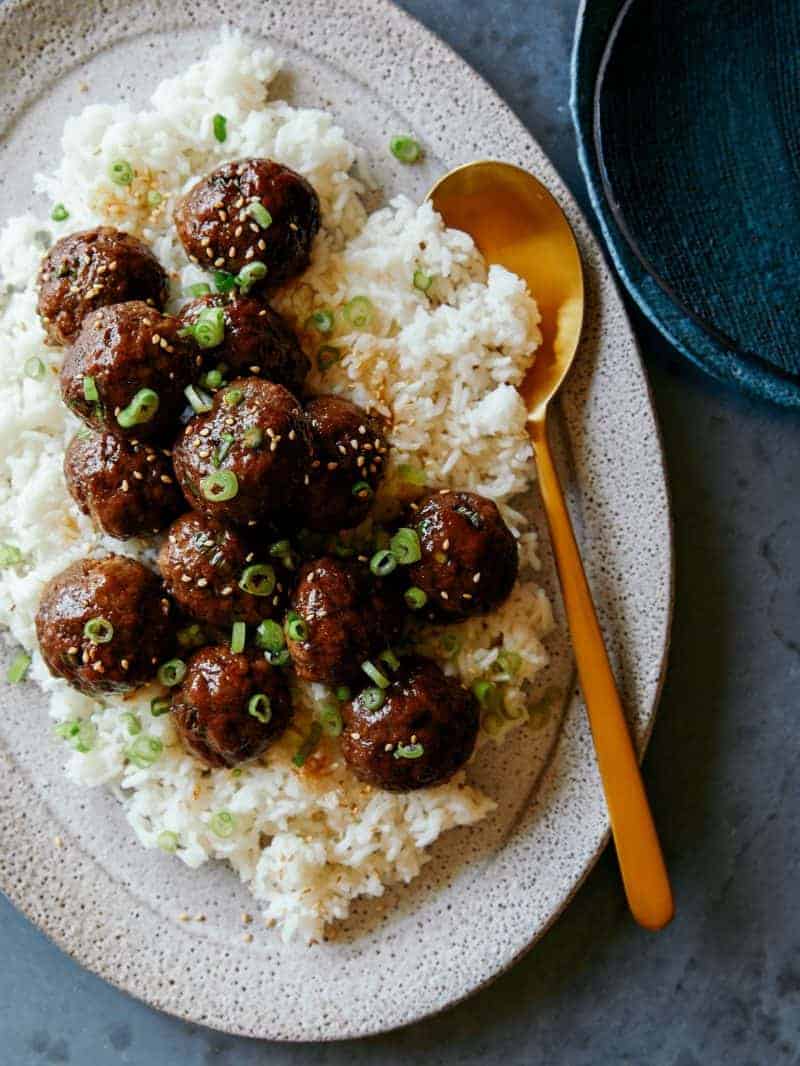 A platter of Korean BBQ meatballs over a bed of rice with plates on the side. 