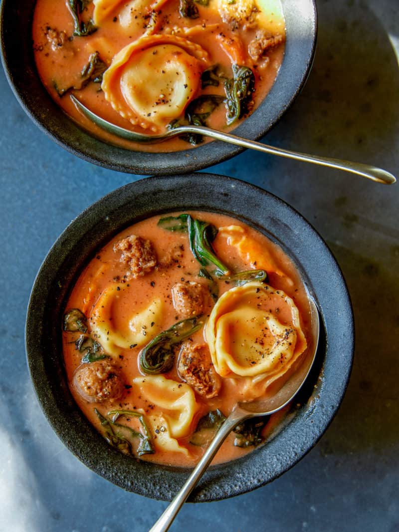 A close up of creamy tortellini with sausage and spinach soup in blue bowls with spoons.