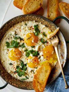 A pan of herb baked eggs with crumbled cheese with bread and a fork.