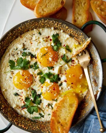 A pan of herb baked eggs with crumbled cheese with bread and a fork.