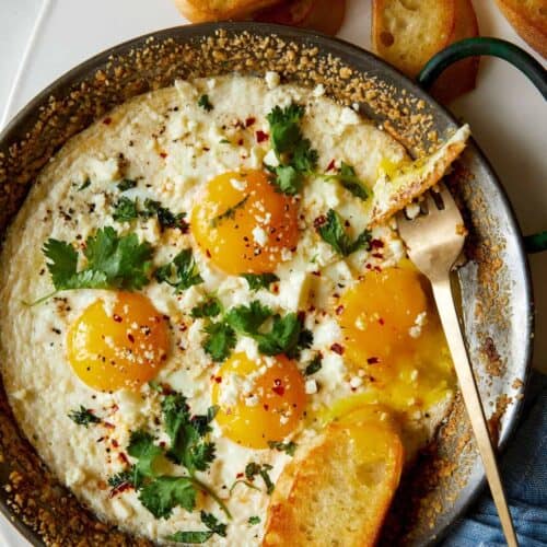 A pan of herb baked eggs with crumbled cheese with bread and a fork.