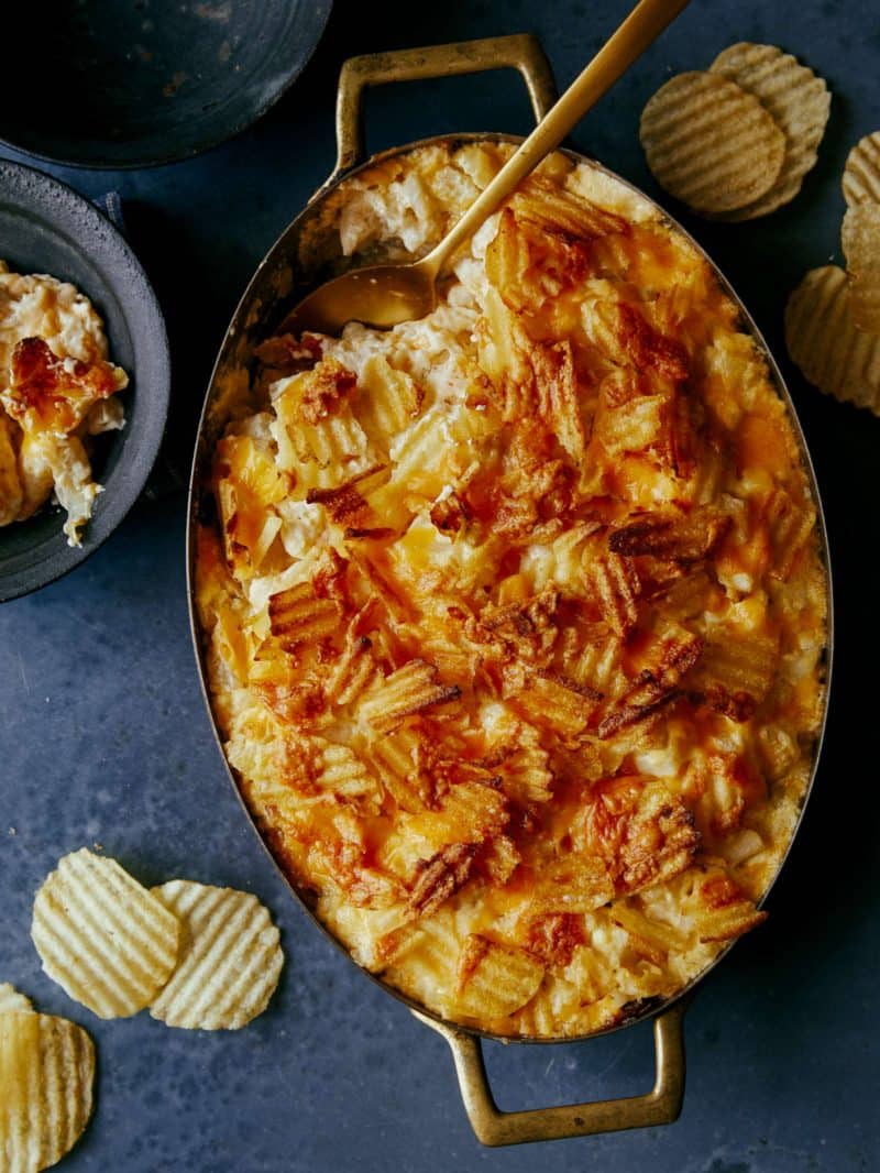 Cheesy potatoes with a potato chip crust and caramelized onions with a scoop out. Potato chip topping scattered around the side and some bowls ready for potatoes. 