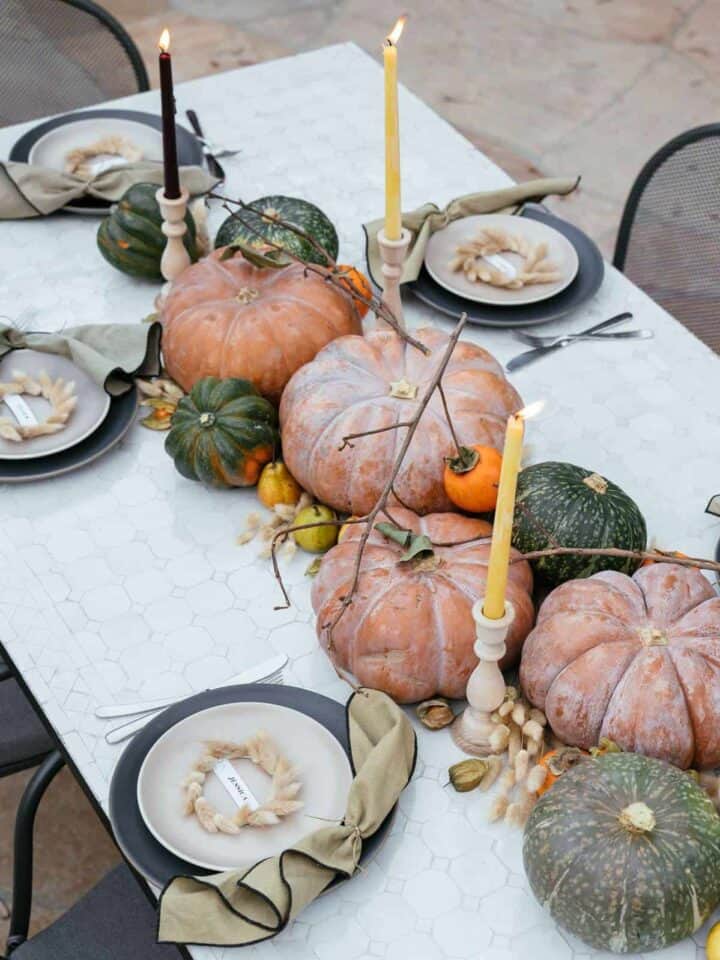 A table with place settings, winter squash, fruits, dried bunny tails, and candles in center.