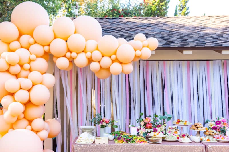 Balloon garland surrounds a table of food with streamer backdrop.