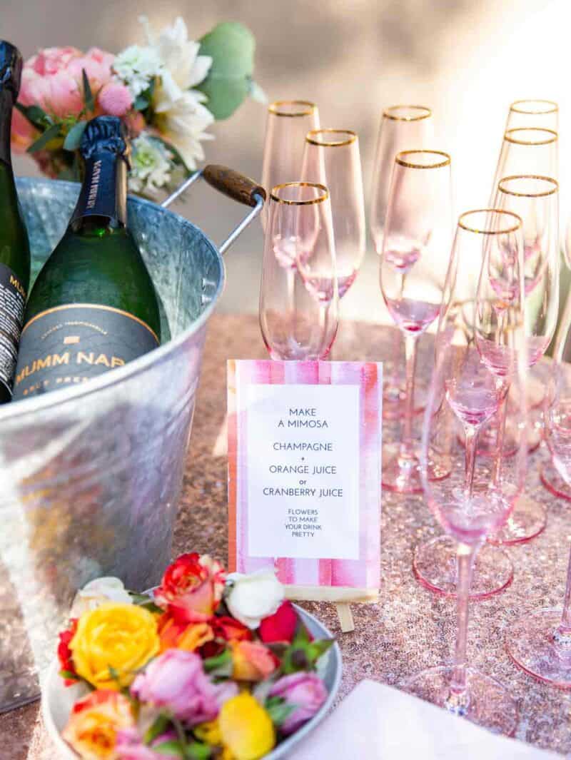 Mimosa bar with champagne bottles, pink glasses, and a sign.