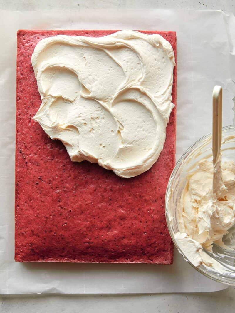 A whole raspberry sheet cake, half frosted with frosting in a bowl on the side.