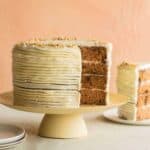 A recipe for Carrot Cake with Cream Cheese Frosting on a pedestal with a slice taken out, a plate to the right with the slice on it. The cake is three layers.