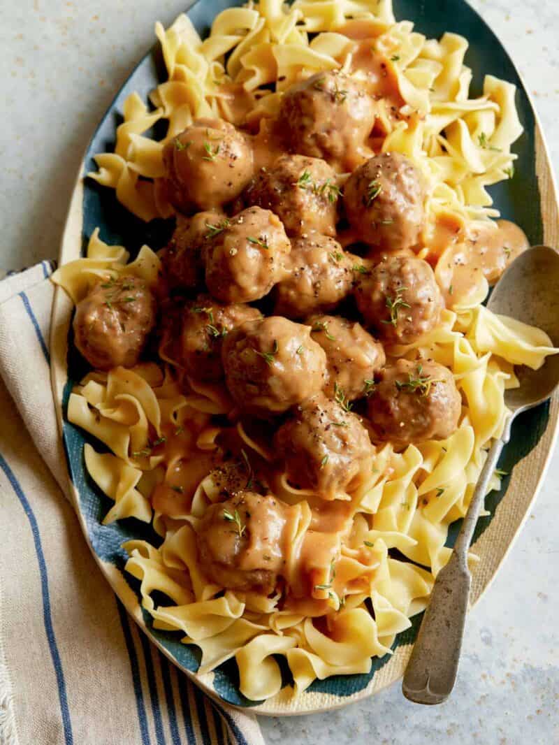 A platter of lamb meatballs covered in gravy on top of egg noodles with a spoon.