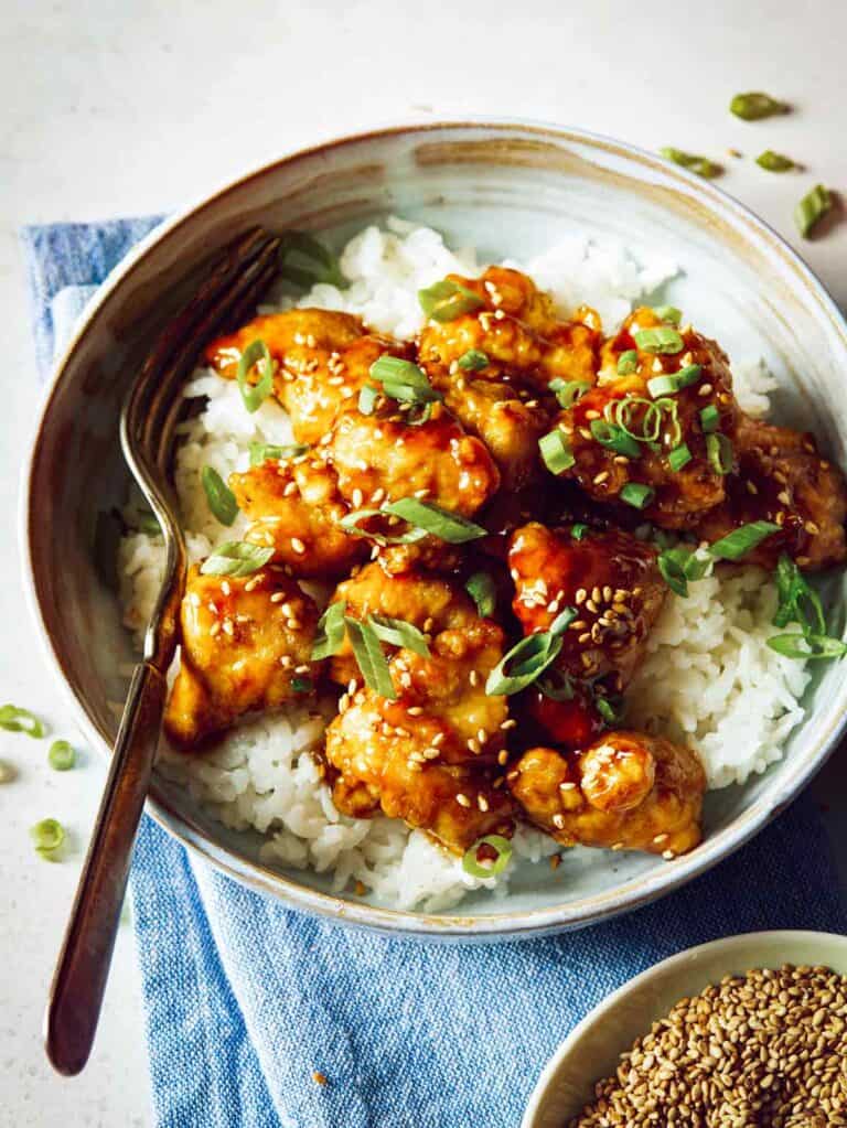 A close up of sesame chicken on white rice in a bowl with a fork.