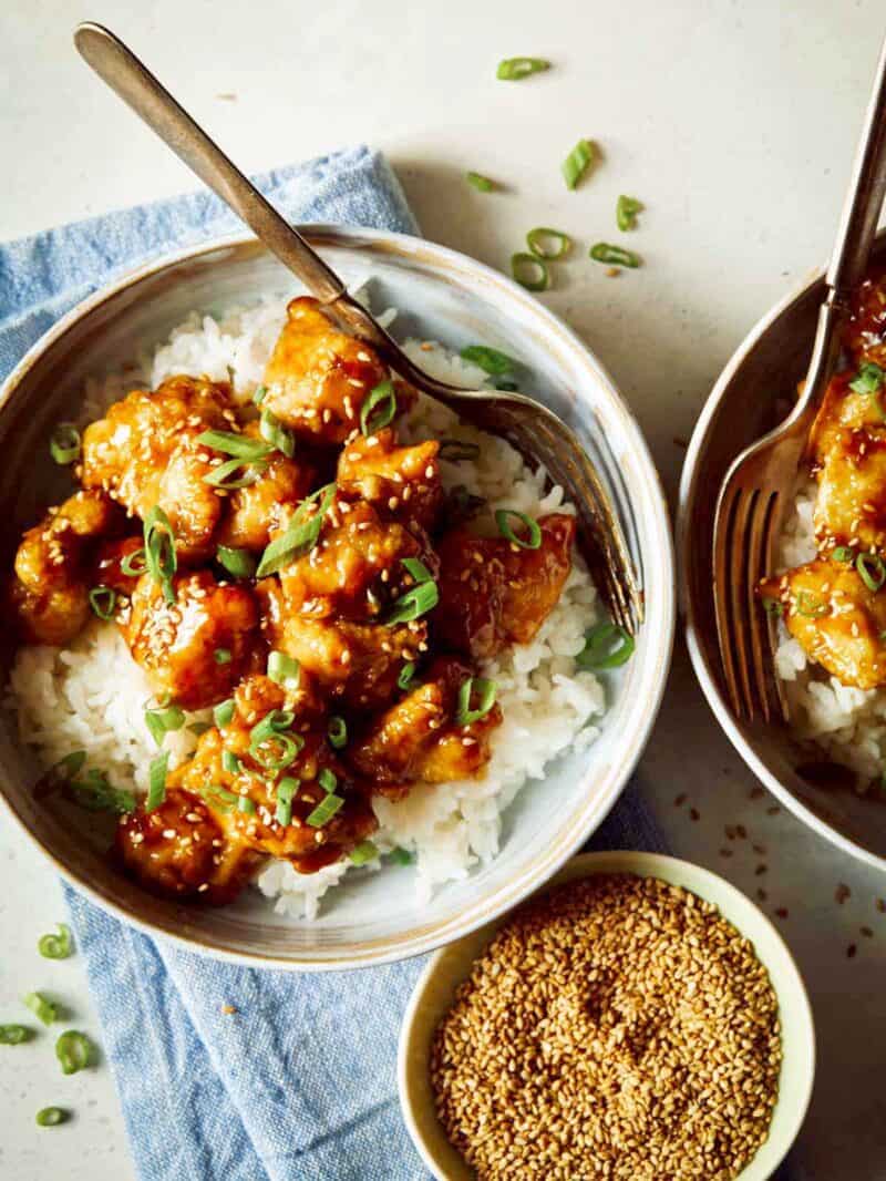 Two bowls of fresh sesame chicken in bowls with rice and a little bowl of sesame seeds.