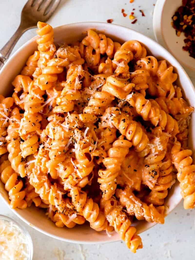 heaping bowl of finished rotini tossed in vodka sauce, topped with Parmesan and black pepper with fork off to side and small bowls of crushed red pepper flakes and more grated Parmesan