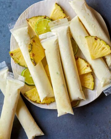 A plate of pina colada Italian ice pops with fresh pineapple and lime slices.