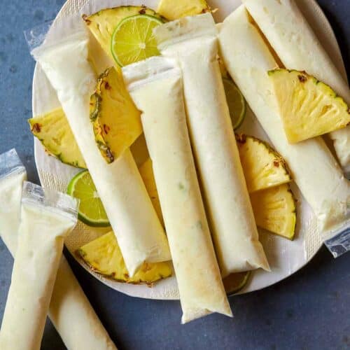 A plate of pina colada Italian ice pops with fresh pineapple and lime slices.