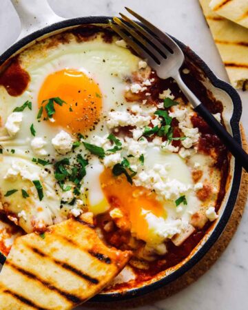An individual shakshuka with hominy and feta and a fork.