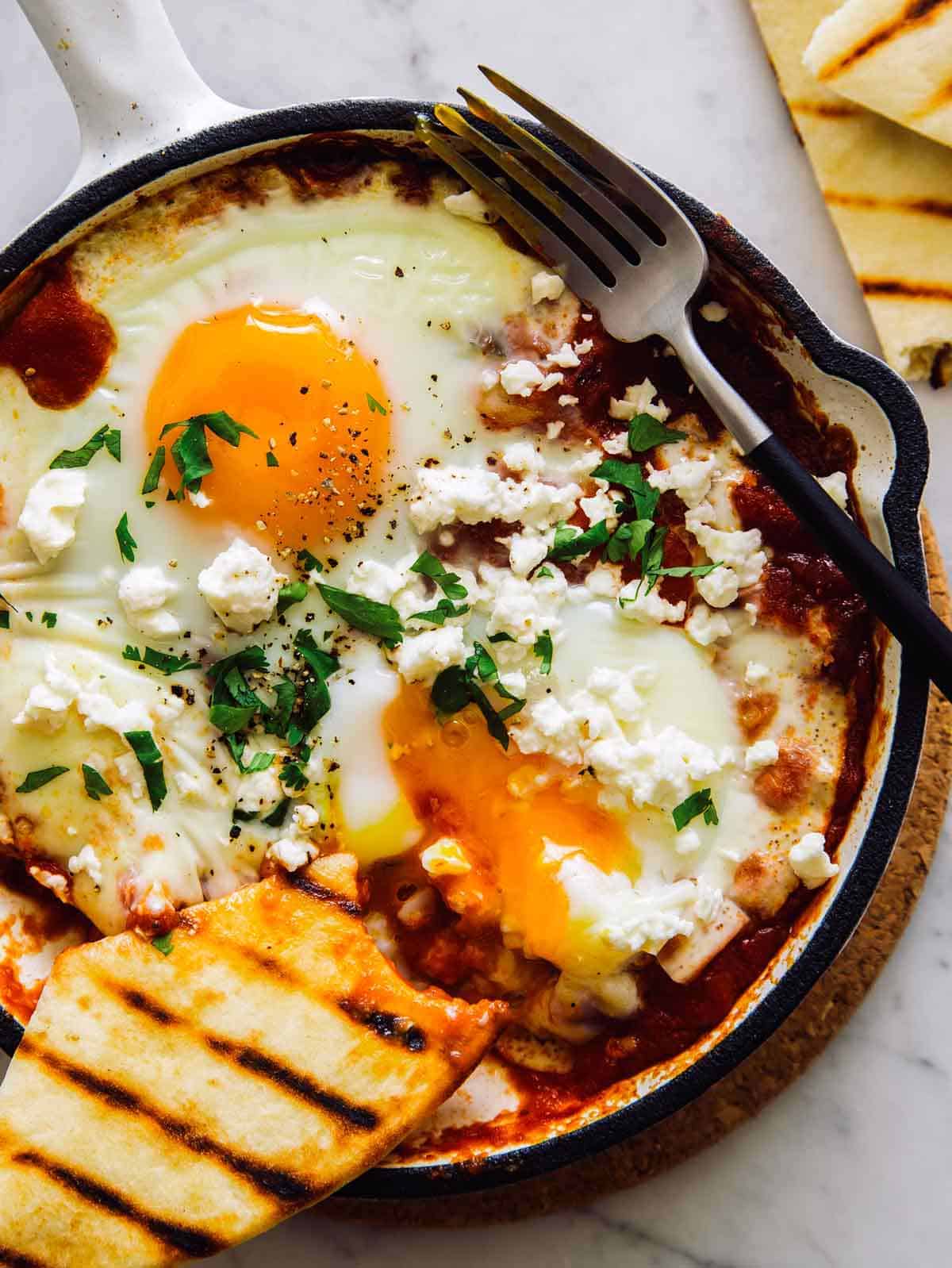 Individual shakshuka with hominy and feta, with a broken egg yolk and some crusty bread. 