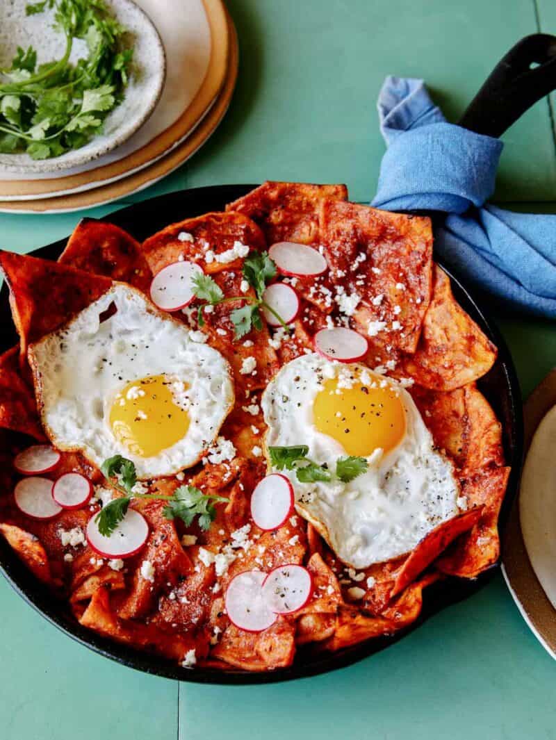 Chilaquiles with two fried eggs on top in skillet. Bowl of cilantro sprigs above skillet