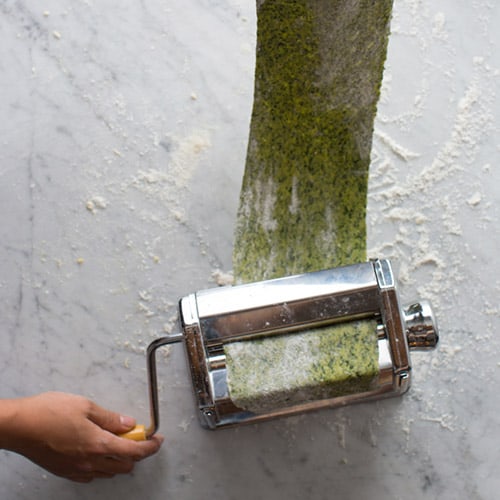 Large green sheet of pasta running through a pasta roller with a hand on the crank.