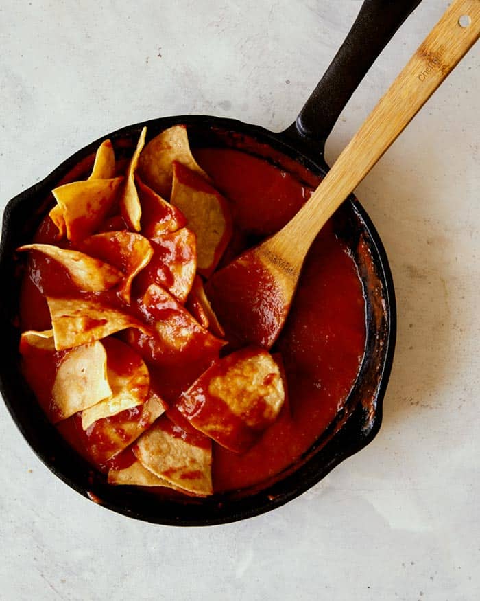 Tortilla chips in a skillet being gently folded together with a red sauce to make chilaquiles.