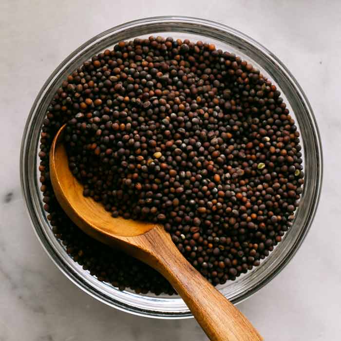 A glass jar full of brown mustard seeds and a wooden spoon in the seeds. 