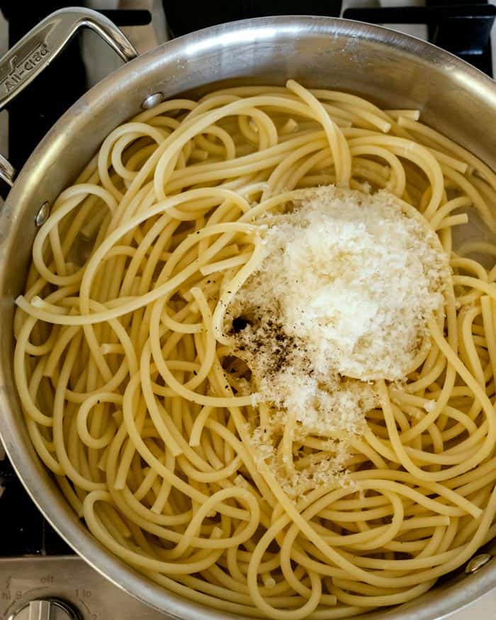 Cooked bucatini noodles in a skillet with grated cheese and cracked black pepper.