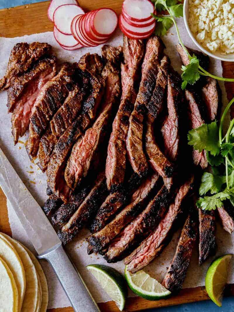 Sliced carne asada with a knife, tortillas, cilantro and radishes.