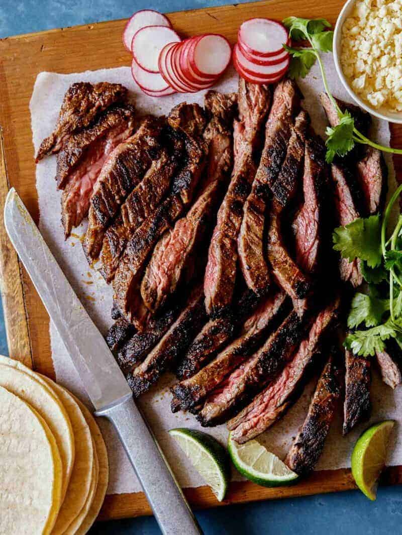 Freshly grilled and sliced Carne Asada on a cutting board with some taco fixings around it. 