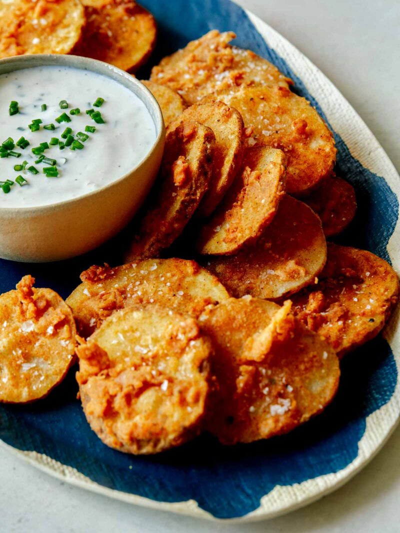 A platter of chicken fried potato slices with a bowl of ranch dressing on the plate.