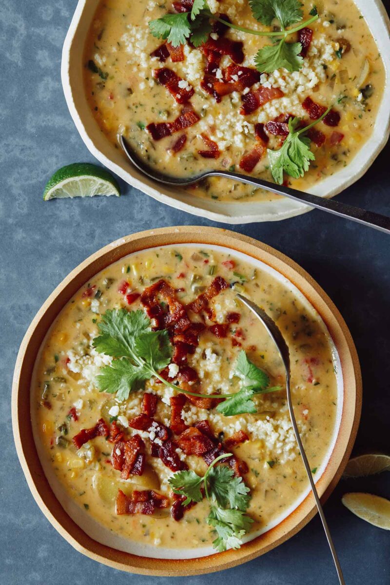 Bowls of corn chowder garnished with bacon, cilantro, cotija, and lime wedges with spoons. 