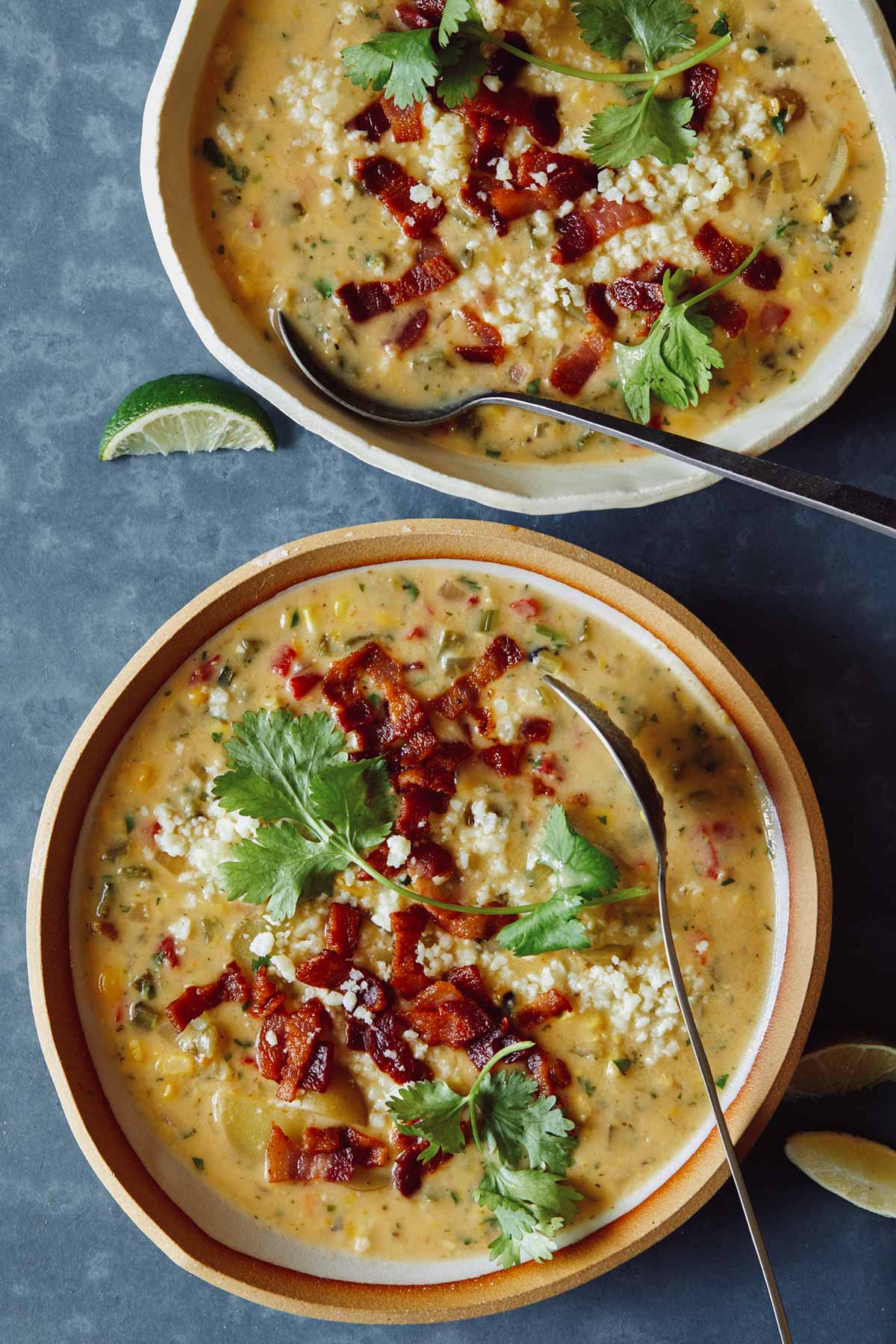Two bowls of corn chowder overhead with some lime wedges on the side and spoons.