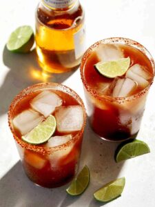 Beer bottle and glasses of Michelada with lime wedge garnish.