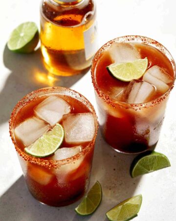 Beer bottle and glasses of Michelada with lime wedge garnish.
