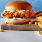 A close up of Popeyes Chicken Sandwich with fries on a wooden board.