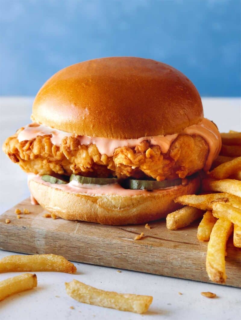 A close up of a Popeyes chicken sandwich with fries on a wooden board.
