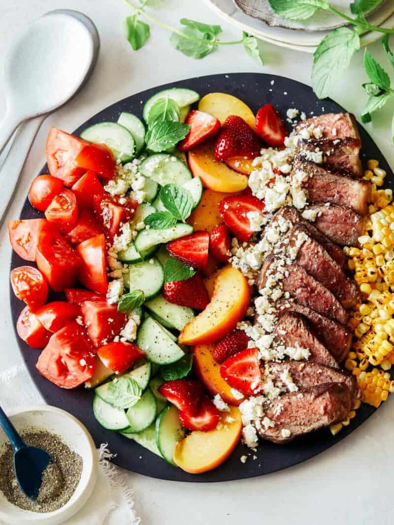 A summer steak salad on a platter with fresh herbs on the side and a stack of plates. 