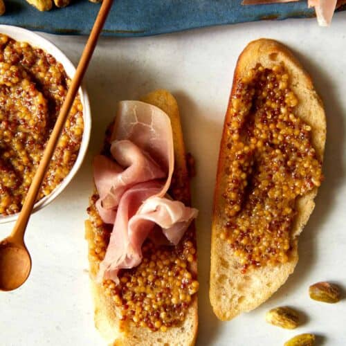 A bowl of whole grain mustard with a spoon and spread on bread.