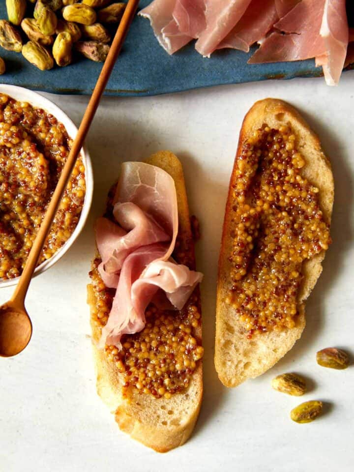 A bowl of whole grain mustard with a spoon and spread on bread.