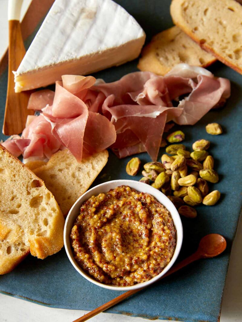 Whole Grain Mustard in a bowl on a plate with cheese and prosciutto. 