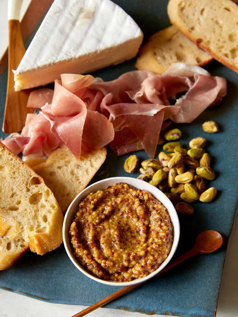 Whole Grain Mustard in a bowl on a plate with cheese and prosciutto.