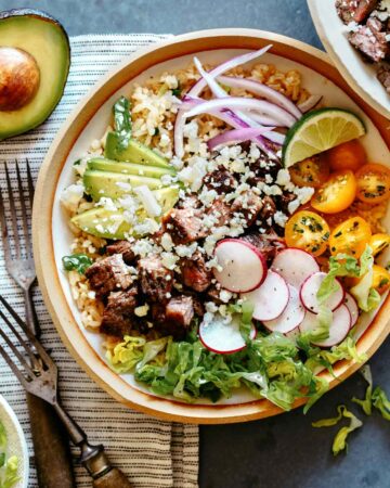 A Carne Asada Burrito bowl with forks on the side and half an avocado.