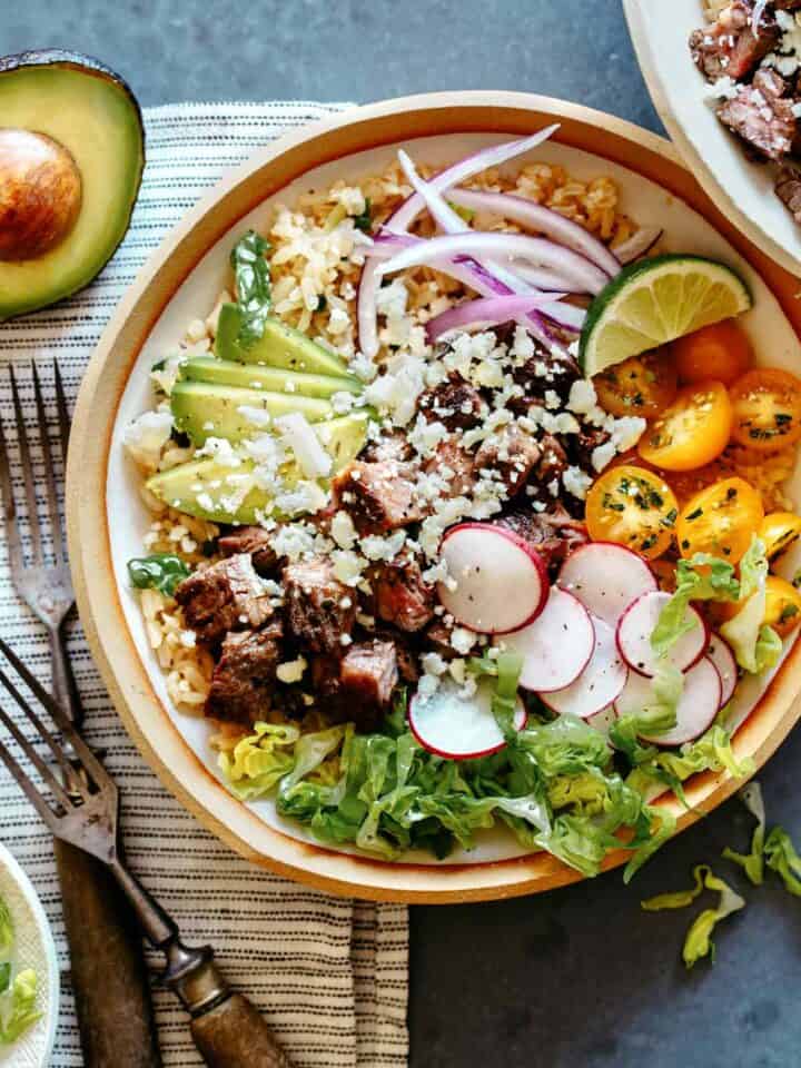 A Carne Asada Burrito bowl with forks on the side and half an avocado.