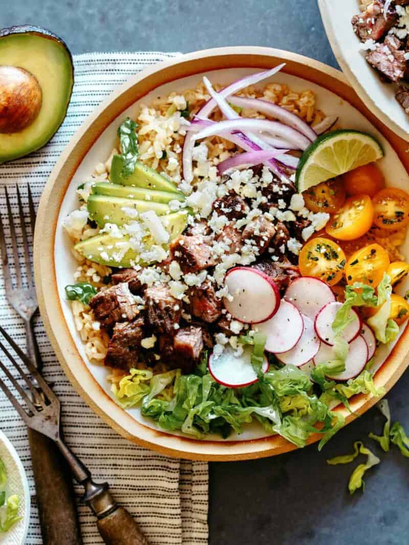 A Carne Asada Burrito bowl with forks on the side and half an avocado. 