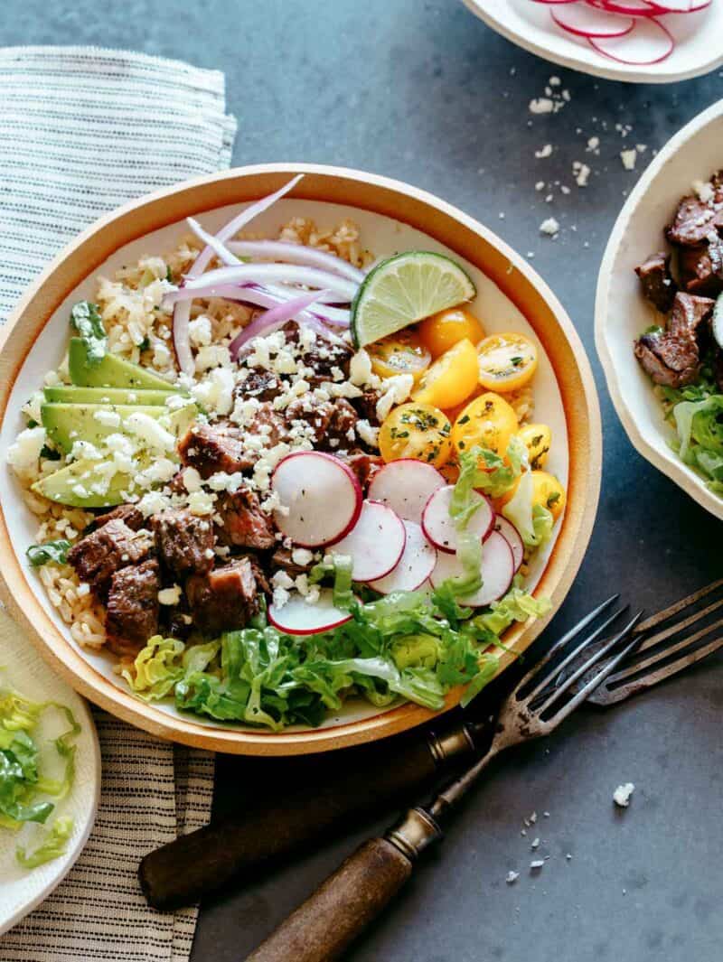 Two bowls of carne asada burrito bowls with toppings peeking in and forks on the side. 