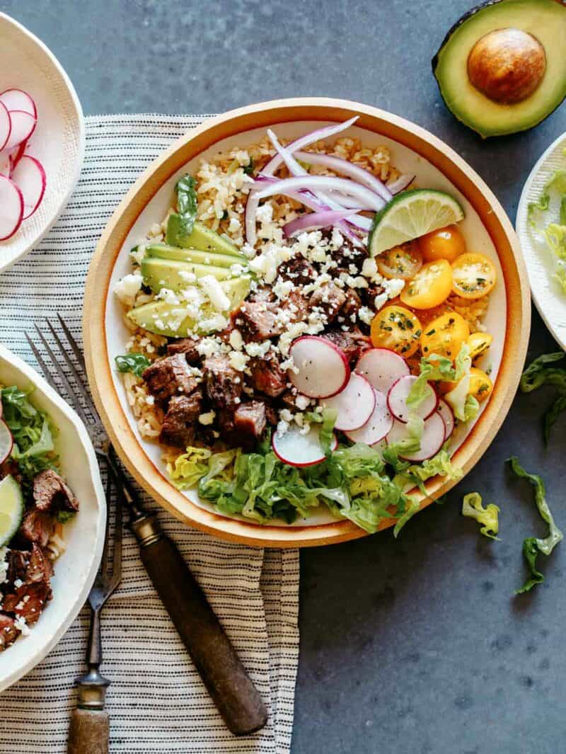 Carne Asada Burrito bowl with a lot of toppings, and avocado on the side, with another bowl peeking in. 