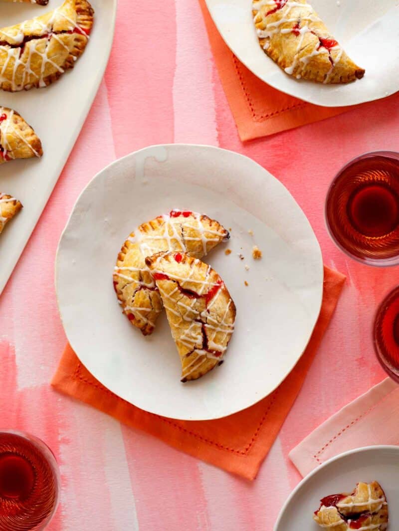 Cherry hand pies with cherry icing drizzle on plates next to drinks.
